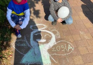 Anioł narysowany kredą na chodniku. Dwie dziewczynki siedzą obok anioła. Na skrzydłach dziewczynki wpisały swoje imiona Oliwia i Zosia.